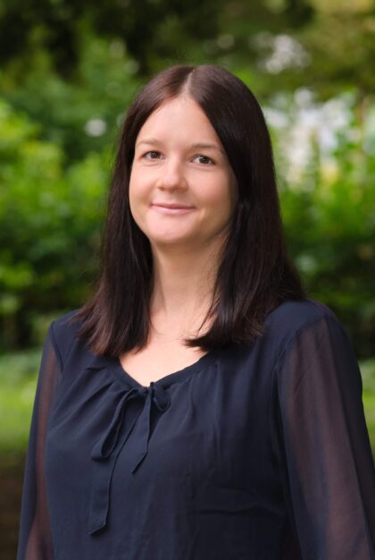 Frau Bruss steht vor ein paar Bäumen und blickt in die Kamera.