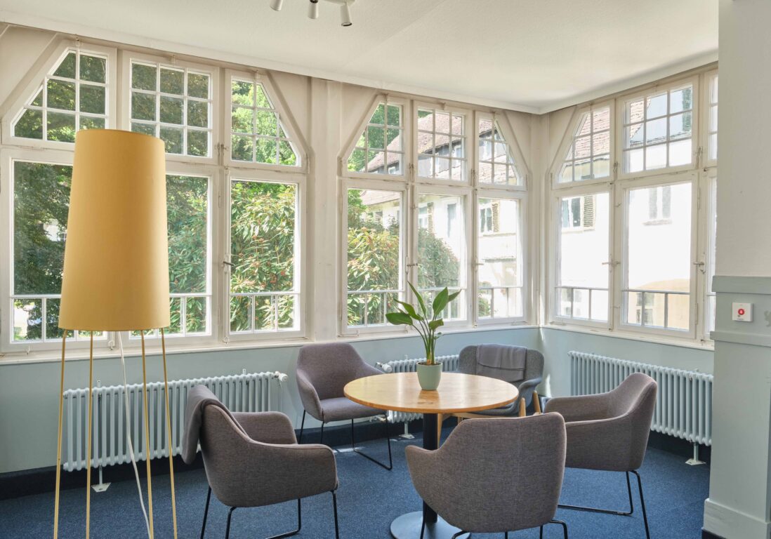 Ein heller Raum ist zu sehen. Eine Lampe steht neben einem Holztisch mit 4 bequemen Stühlen.