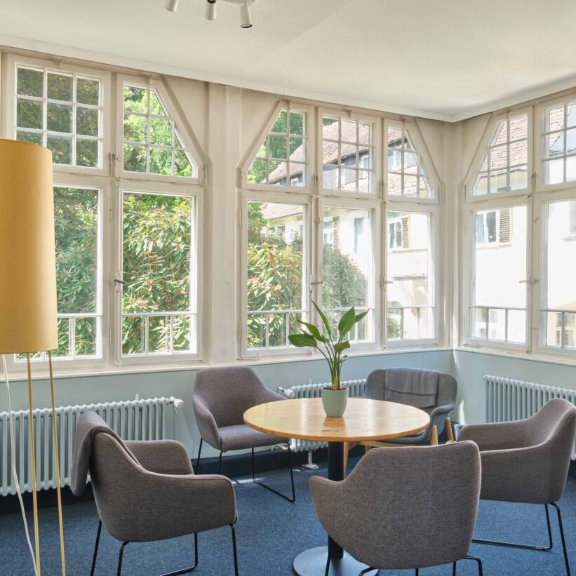 Ein heller Raum ist zu sehen. Eine Lampe steht neben einem Holztisch mit 4 bequemen Stühlen.